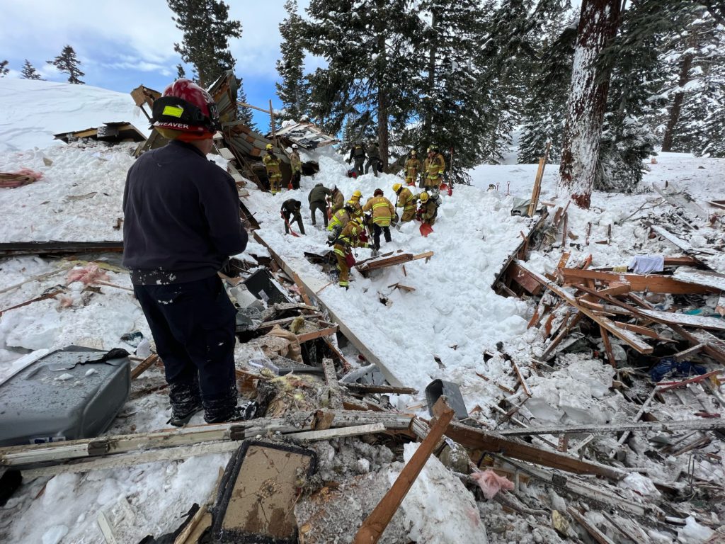 Fire crews search an exploded home in the snow. One member stands above the rest in a blue uniform and helmet.
