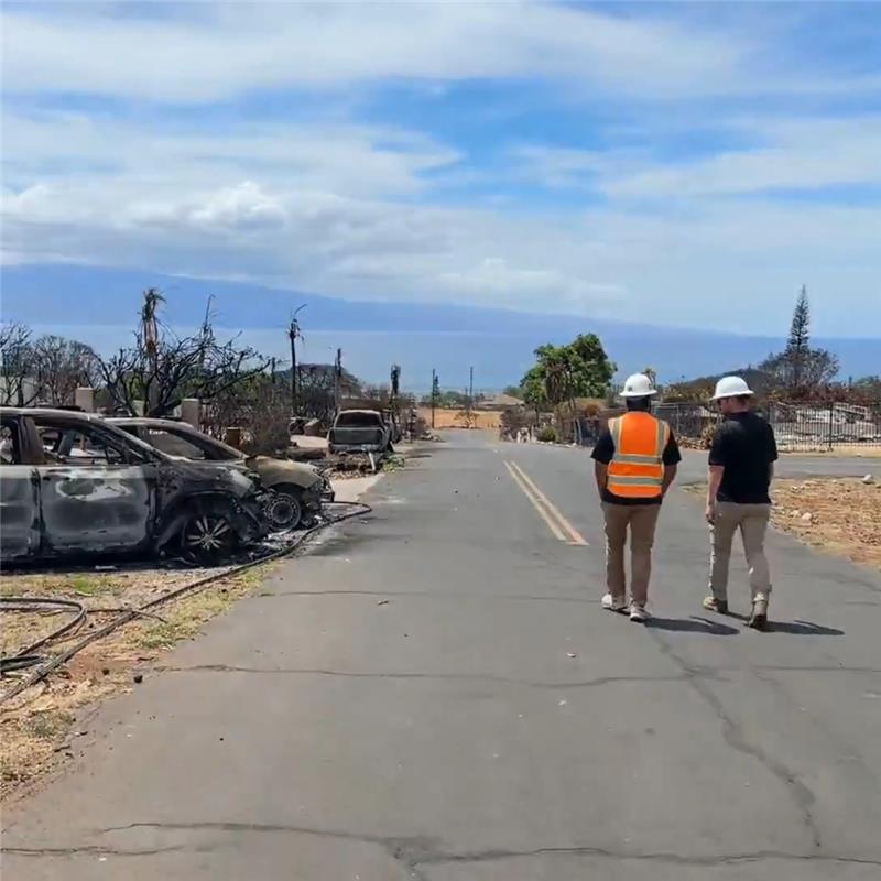 3a28cb6c-428a-467a-ada2-7cf1c33f79b2 Two Cal OES employees walk along a road on Maui. The impacts of the Lahaina wildfire, including burned vehicles, are visible around them.