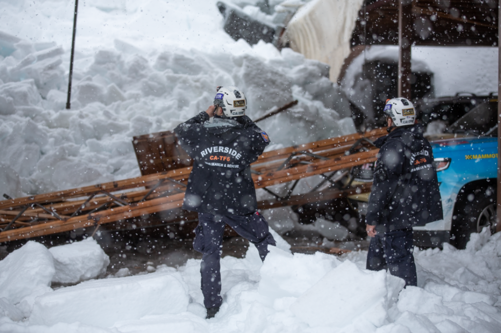 Two individuals stand in the snow next to a collapsed structure. They both wear jackets that read "RIVERSIDE CA-TF6 URBAN SEARCH & RESCUE"