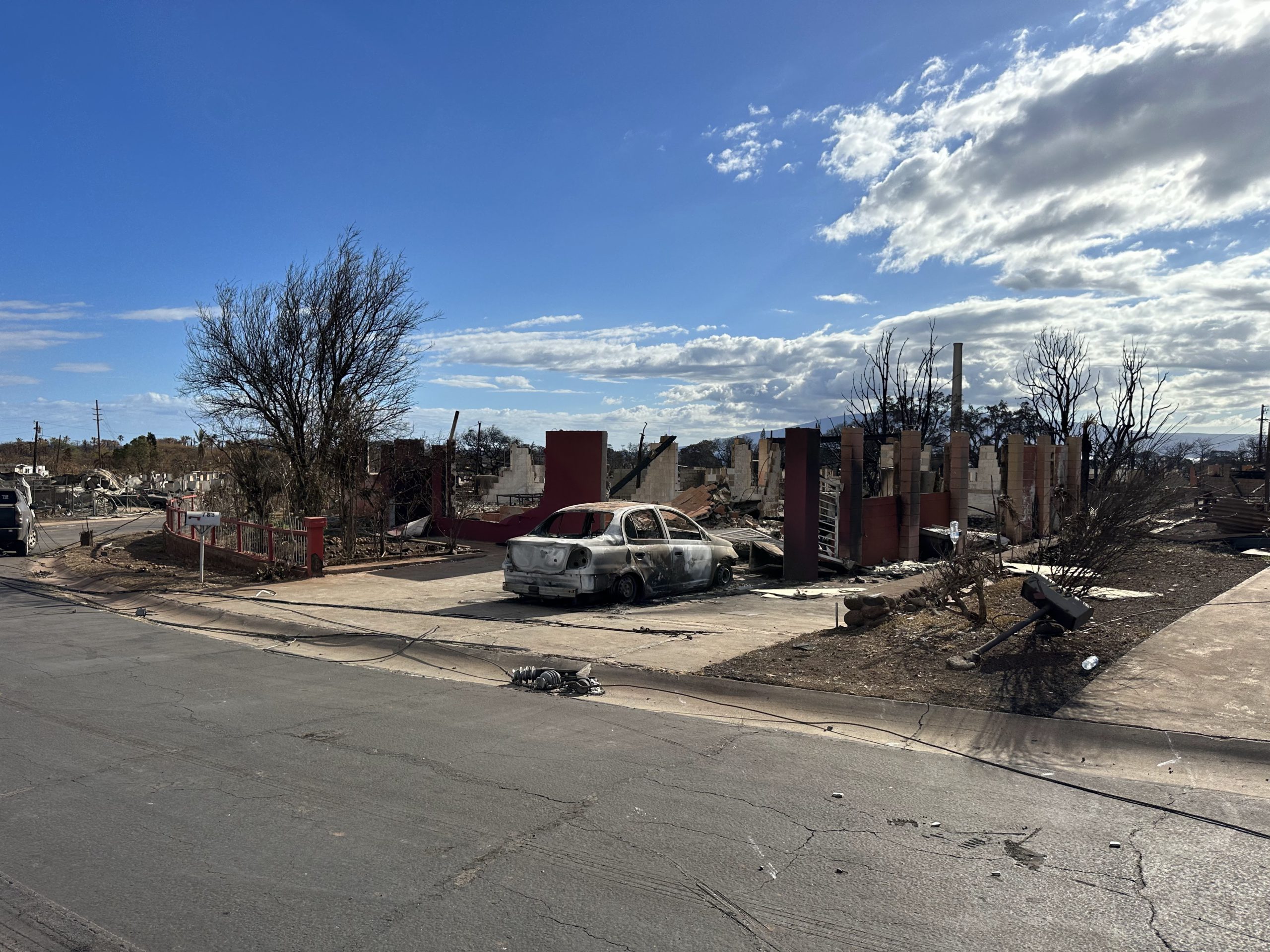A burnt-out car in a driveway in Lahaina, Hawaii. Behind it lies remnants of Lahaina, Hawaii. 