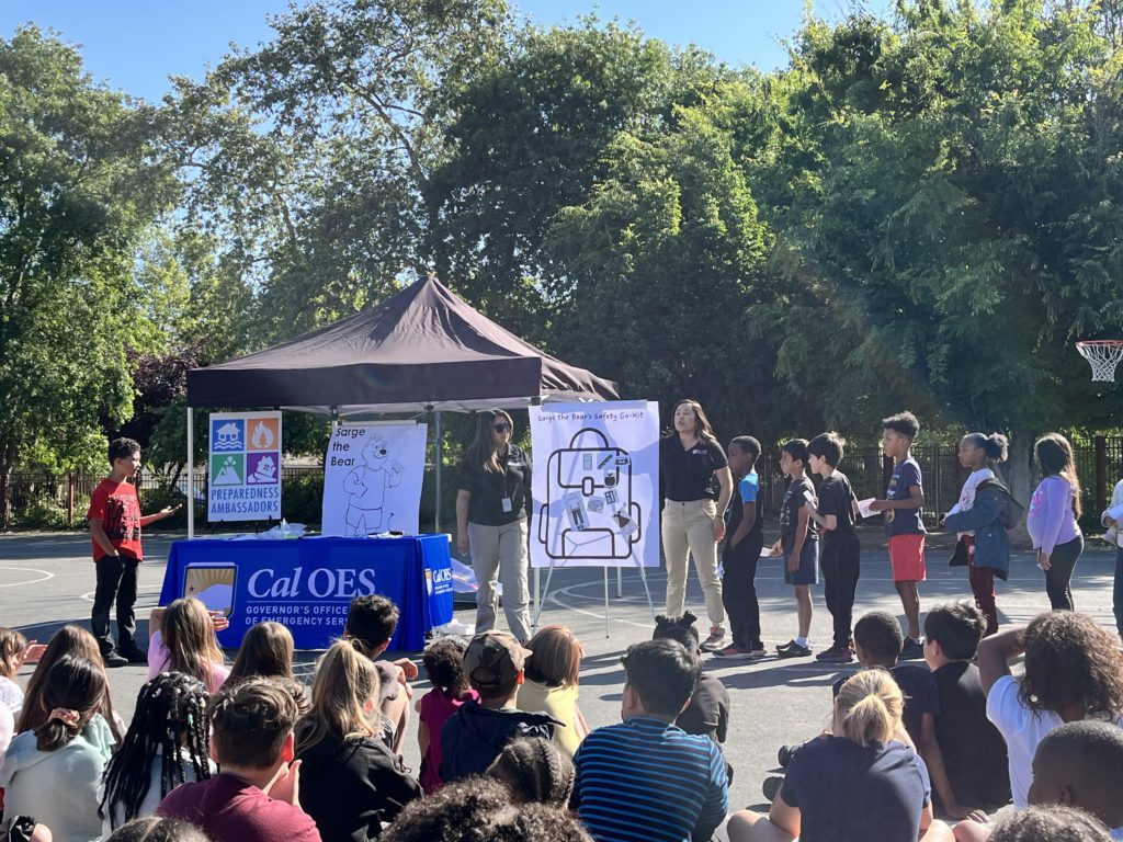 group of students watch sit on a blacktop watching two Cal O E S representatives in front of a Cal O E S table and pop up tent.