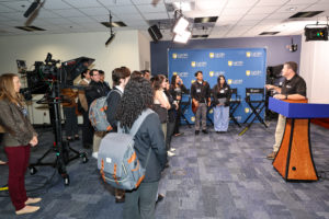 A group of men and women stand in front of a television camera, listening to a man wearing a black polo shirt and tan pants leaning on a podium.
