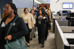 Two women talk to each other as they walk side by side, with people walking ahead of and behind them through an office with cubicles.