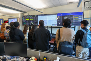 A group of people with their backs to the camera and facing multiple video screens listen to a woman facing them.