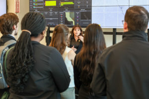 A woman with large video screens behind her talks to a group of people.